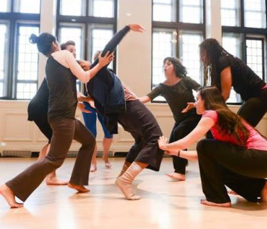 A group of barefoot dancers supporting one another during an expressive healing dance workshop in a bright studio.
