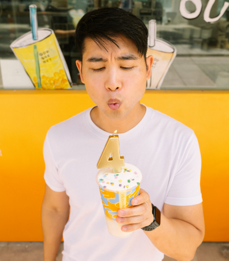 Photo of a young man holding a boba cup with a number 4 candle on top, blowing out the candle's flame